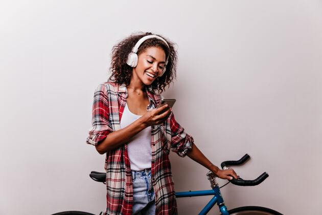 休闲戴着耳机看电话留言的迷人时尚女人穿着考究的非洲女孩拿着自行车拿着智能手机时尚休息活跃