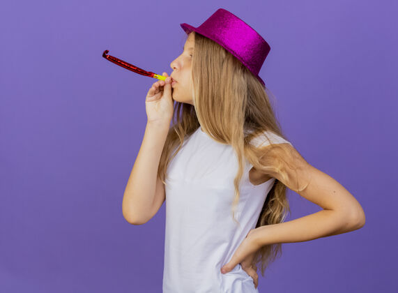 生日美丽的小女孩在节日帽子吹哨子快乐和积极 生日聚会的概念站在紫色的背景正面吹派对
