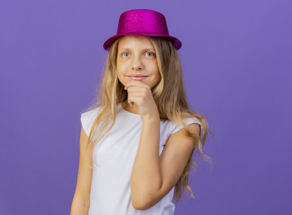 漂亮戴着节日礼帽的漂亮小女孩看着相机 手放在下巴上微笑着 紫色背景下站着生日派对的概念看手小