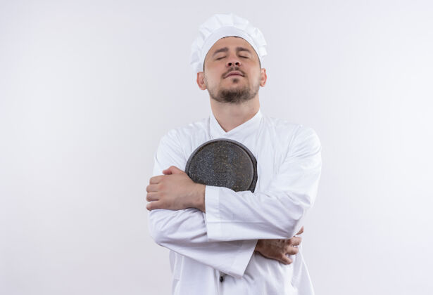 和平穿着厨师制服的年轻帅气的厨师手拿着煎锅 闭着眼睛看着与世隔绝的白色空间帅气持有厨师