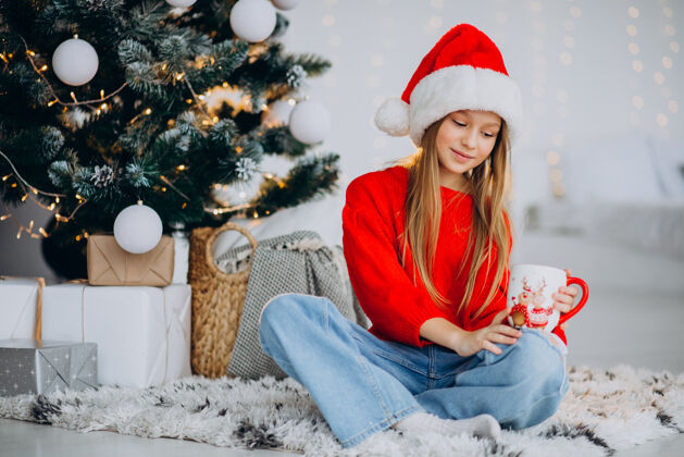微笑在圣诞树旁喝可可的女孩美丽欢呼礼物