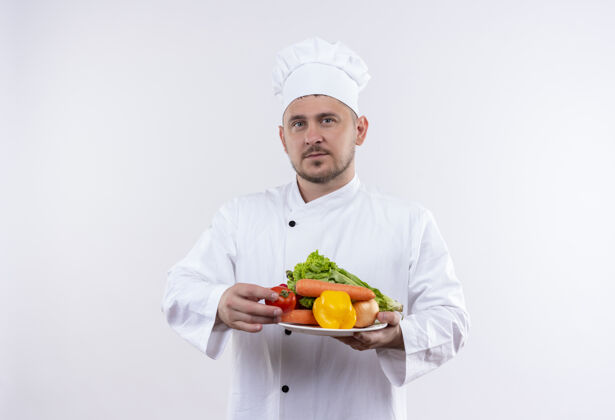 制服年轻帅气的厨师穿着厨师制服拿着盘子里的蔬菜看着孤立的白色空间看着蔬菜厨师