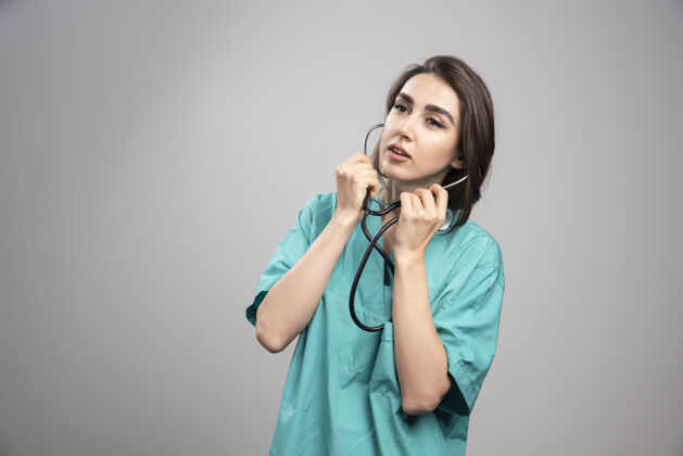 护理年轻的医生穿着制服在灰色的墙上用听诊器成人医疗制服