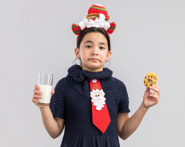 头小女孩穿着针织连衣裙 头上系着一条红色领带 头上戴着一个有趣的圣诞戒指 手里拿着一杯牛奶和饼干 表情悲伤悲伤玻璃杯边