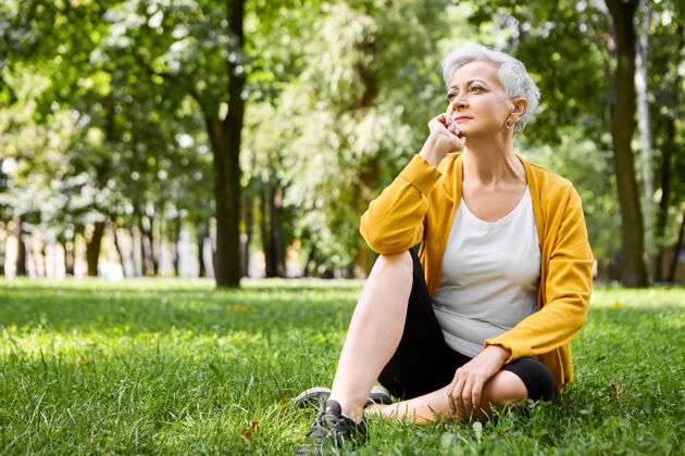 宁静一幅体贴的退休妇女的画像 穿着跑鞋 舒舒服服地坐在绿草上 手放在下巴下 看着人们在公园里散步 神情沉思 心情轻松活跃户外公园
