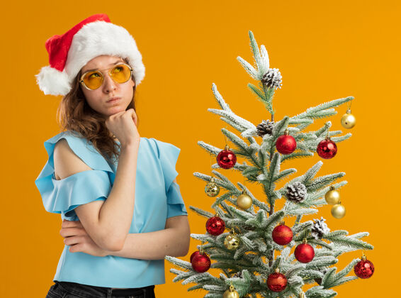 圣诞树身穿蓝色上衣 戴着黄色眼镜的圣诞老人帽的年轻女子站在橙色背景下的圣诞树旁 困惑地抬起头来帽子圣诞老人橙色