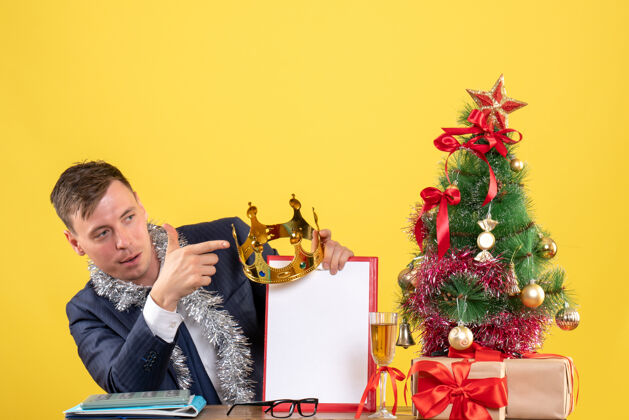 树商务人士手持皇冠和剪贴板坐在圣诞树旁的桌子前 黄色的礼物坐皇冠礼物