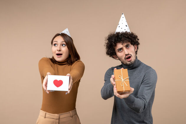 模特惊讶的年轻夫妇戴着新年礼帽摆姿势 镜头上的女孩展示心形 而男人则是灰色的礼物女士情侣帽子