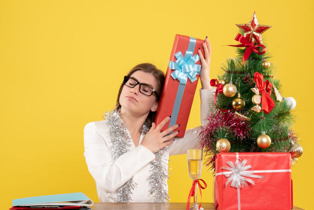漂亮正面图：女医生坐在桌子前 拿着礼物 黄色背景上的圣诞树和礼盒女性健康树