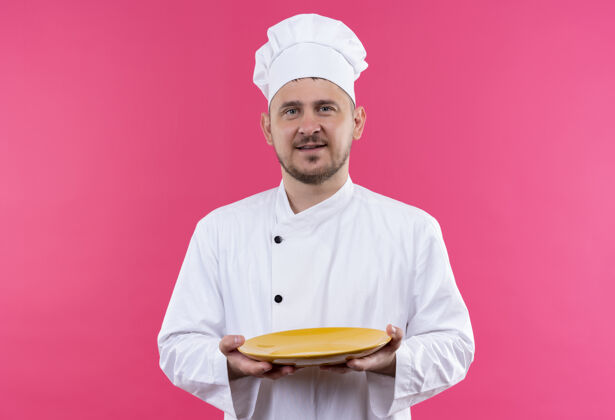 年轻高兴的年轻帅哥厨师穿着厨师制服拿着盘子隔离在粉红色的空间里粉色烹饪手持