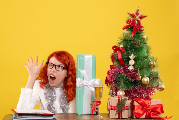 圣诞节前视图：女医生拿着圣诞礼物和黄色背景上的圣诞树坐着坐快乐树