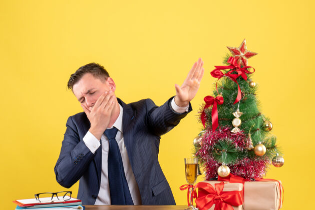 桌子前视图中疲惫的男子打哈欠 而坐在桌旁圣诞树和礼物上的黄色成年人而演讲者