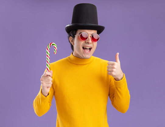 展示身穿黄色高领毛衣 戴着黑帽子 手持糖果手杖 高高兴兴地竖起大拇指的年轻人站在紫色的墙上穿着高领毛衣站着