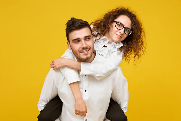 男朋友戴着眼镜的快乐的年轻黑发女人的肖像 骑在男友的背上 靠着空白的黄色墙壁爱 浪漫 团结和幸福的概念肖像帅哥女人