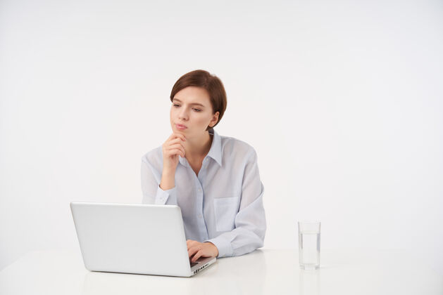 办公室沉思的年轻漂亮的棕色头发的女人 一头时髦的短发叠着嘴唇 一边沉思地看着她的笔记本电脑 举起手托着下巴 坐在白色的椅子上年轻坐着沉思
