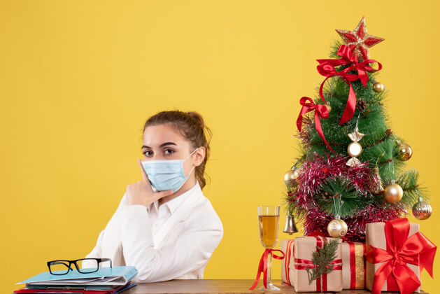 外套正面图女医生戴着黄色背景的无菌口罩坐在圣诞树和礼盒里成人肖像实验室
