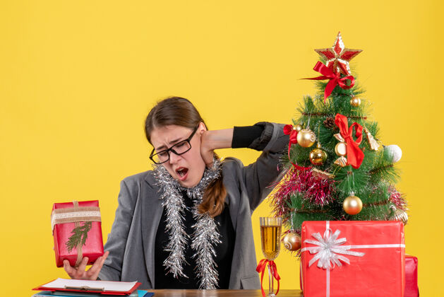 桌子前视图疲惫的女孩戴着眼镜坐在桌旁 抱着她的脖子圣诞树和鸡尾酒礼物累了脖子导演