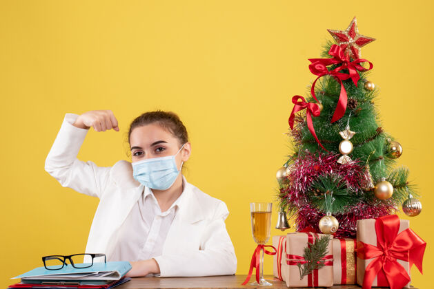实验室外套正面图女医生坐在无菌面具上 在黄色背景上弯曲着圣诞树和礼品盒医生情感弯曲