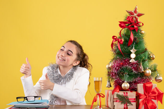 坐着前视图：女医生坐在桌子后面 手里拿着圣诞礼物和树 在黄色背景上和某人说话前面谈话新的