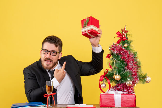 生活正面图：年轻的男工人在桌子后面拿着礼物 圣诞树挂在黄色的树上工人年轻人男