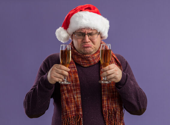 圣诞快乐困惑的成年男子戴着眼镜 戴着圣诞帽 脖子上围着围巾 手里拿着两杯香槟 看着一个孤立的紫色背景眼镜快乐抱着