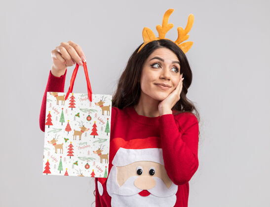 看高兴的年轻漂亮女孩戴着驯鹿鹿角头带和圣诞老人毛衣拿着圣诞礼品袋手放在脸上看着身边年毛衣抱