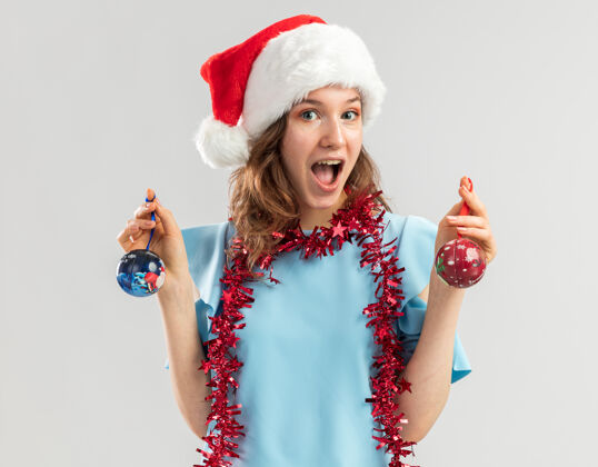 新一位身穿蓝色上衣 头戴圣诞帽 脖子上系着金属丝 手持圣诞球的快乐而兴奋的年轻女子 脸上洋溢着欢快的笑容脖子高兴圣诞老人