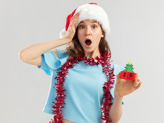 举行身穿蓝色上衣 脖子上戴着镀金圣诞帽的年轻女子手里拿着玩具立方体 手放在头上 看起来很惊奇帽子圣诞快乐