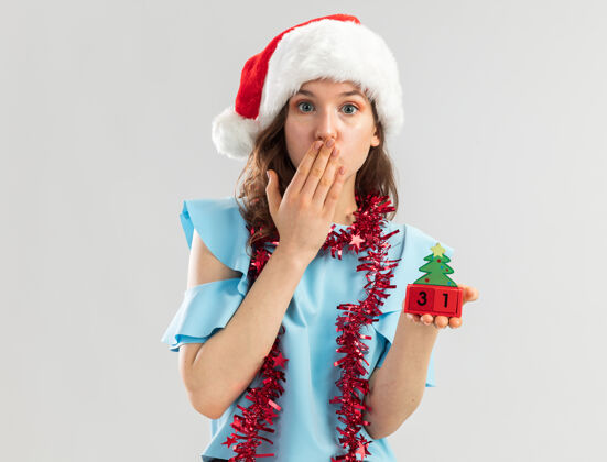 新年穿着蓝色上衣 戴着圣诞帽 脖子上戴着金属丝的年轻女子 手里拿着玩具立方体 看起来像是在用手捂住嘴巴嘴年被