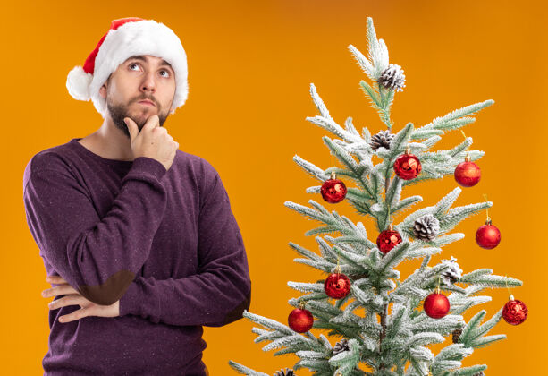 年轻穿着紫色毛衣 戴着圣诞帽的年轻人抬起头 脸上带着沉思的表情 想着橙色背景下的圣诞树表情沉思男人