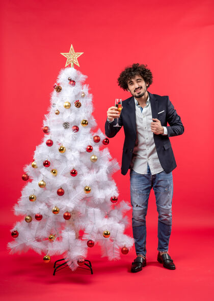 圣诞树圣诞节庆祝活动与留胡子的年轻人与葡萄酒看着相机 站在圣诞树附近的红色年轻人庆祝男人