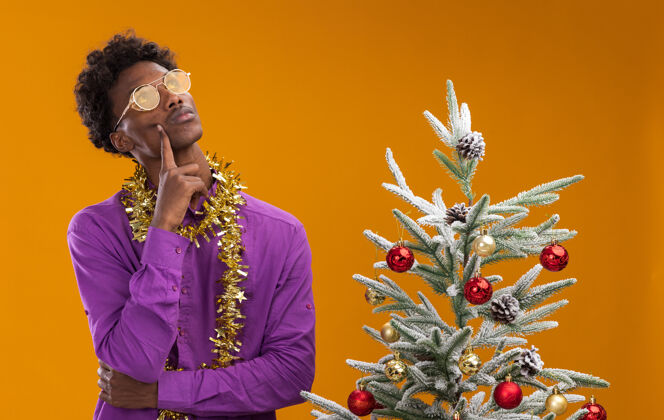 圣诞节体贴的美国黑人年轻人戴着一副眼镜 脖子上戴着金箔花环 站在橙色背景的装饰过的圣诞树旁眼镜装饰圣诞树