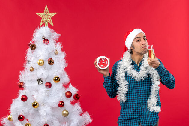 圣诞自信的年轻人戴着圣诞老人的帽子 品尝着一杯葡萄酒 站在圣诞树旁的钟庆典杯子自信