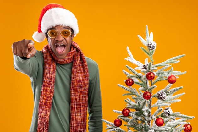 周围戴着圣诞帽 围着围巾 用食指指着镜头的非裔美国人站在橙色背景下的圣诞树旁 既高兴又兴奋圣诞围巾手指