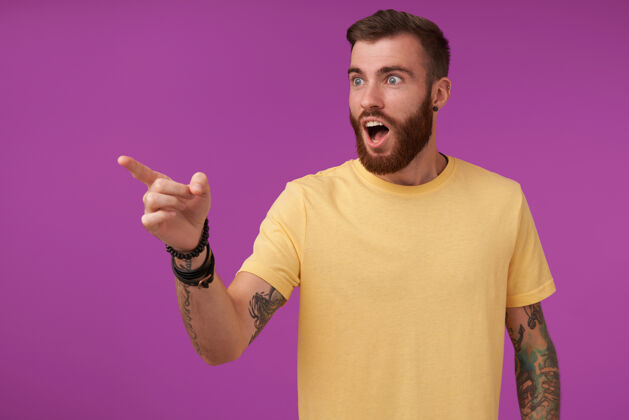 黄色震惊的帅哥 黑发胡子 纹身 穿着黄色t恤和健身手表 注意到一些东西 并用食指在上面显示 孤立的紫色紫色男人身体