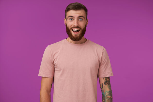 模特一个年轻的留着胡子的黑发男人的肖像 他身上有纹身 穿着米色t恤和时髦的配饰 站在紫色的地面上 双手放下 张大嘴巴米色衣服男性