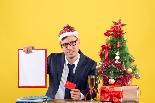 商人前视图的商人持有信用卡和剪贴板坐在圣诞树附近的桌子和黄色的礼物信用办公室持有