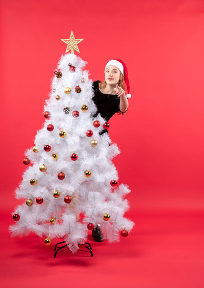 站着新年的气氛与年轻女子在黑色礼服和圣诞老人的帽子站在白色圣诞树后面年轻圣诞老人庆祝