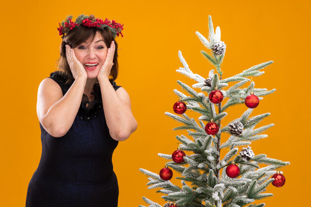 女人兴奋的中年妇女头戴圣诞花环 脖子上戴着金属丝花环 站在装饰好的圣诞树旁头看兴奋