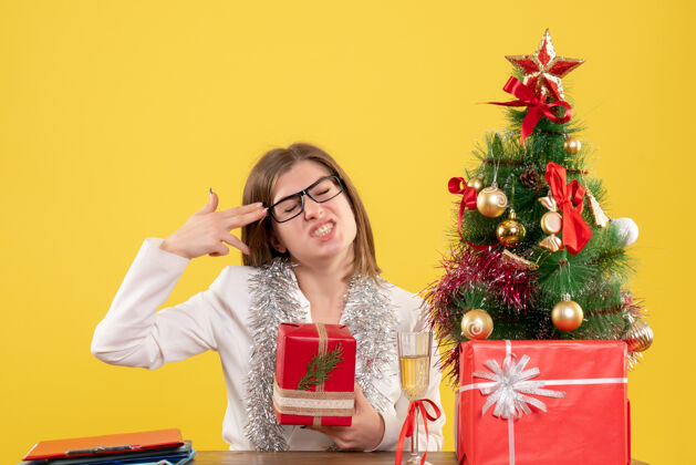 健康正面图：女医生坐在桌子前 拿着礼物 黄色背景上的圣诞树和礼盒人前面背景