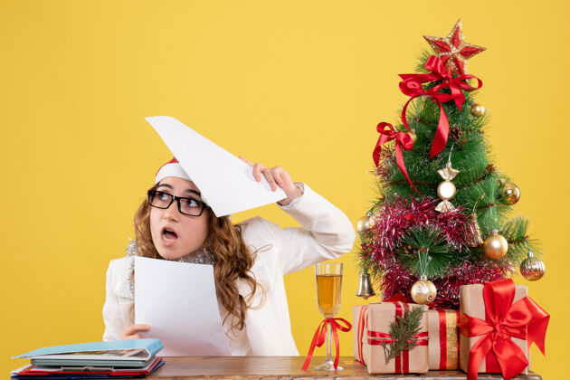 衣服前视图女医生坐在圣诞树上 拿着黄色背景上的文件快乐礼物背景