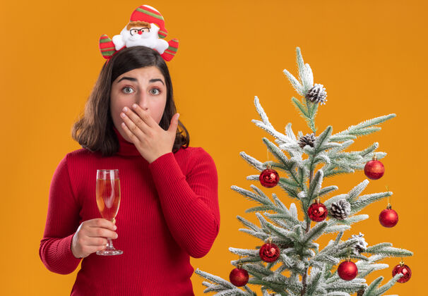 拿着穿着圣诞毛衣的年轻女孩戴着滑稽的头带 拿着一杯香槟放在橙色背景下的圣诞树旁穿被相机