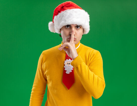 手势一个穿着黄色高领毛衣 戴着圣诞帽 打着滑稽领带的年轻人 严肃地看着镜头 手指放在嘴唇上 在绿色背景上做着沉默的手势搞笑看手指