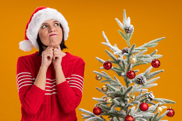 帽子体贴的年轻女孩 戴着圣诞帽 站在装饰好的圣诞树旁 抬起头 双手放在下巴下 噘着嘴唇 孤立地站在橙色的墙上戴着的下巴噘着的
