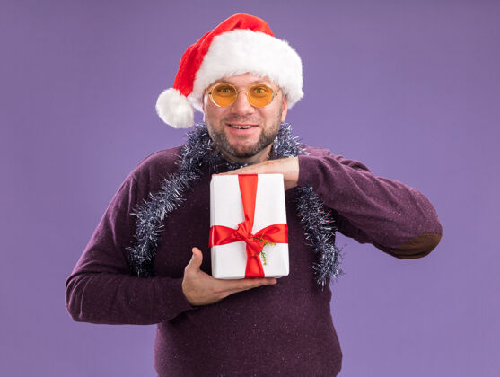 帽子微笑的中年男子戴着圣诞帽 脖子上戴着金箔花环 戴着眼镜 手里拿着礼包 隔离在紫色的墙上金属丝中年男人