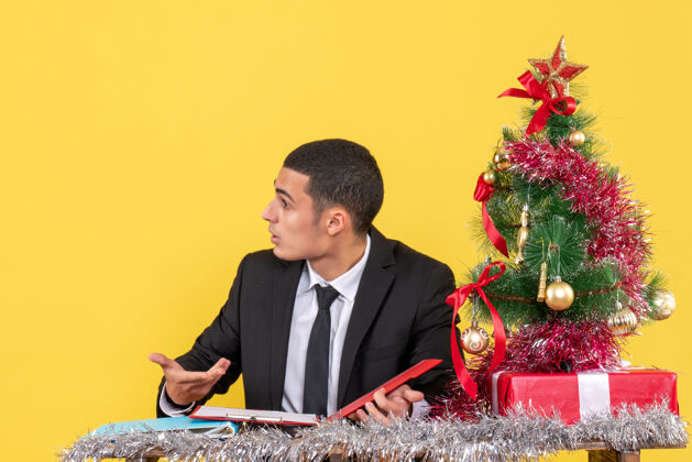 树前视图困惑的西服男子坐在桌子旁拿着文件 看着右边的圣诞树和礼物右男人会议