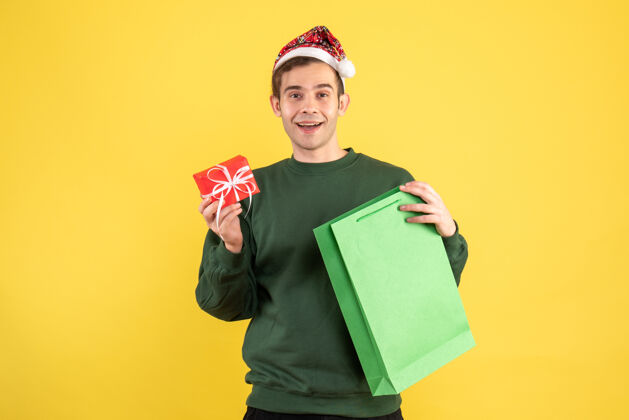 年轻正面图快乐的年轻人戴着圣诞帽 拿着绿色的购物袋和礼物站在黄色的地板上包快乐礼物