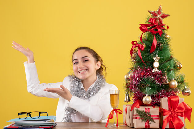 插花正面图：女医生坐在黄色背景的桌子后面 还有圣诞树和礼品盒微笑插花快乐
