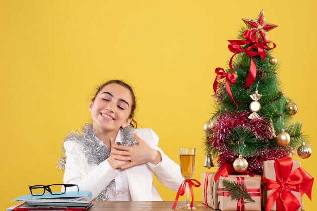 人正面图：坐在桌子后面的女医生在黄色背景上高兴地看着圣诞树和礼品盒高兴人护士