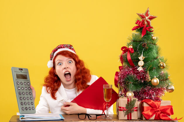 礼物前视图女医生坐在桌子后面拿着礼物拿着计算器 背景是黄色的圣诞树和礼盒计算器医生微笑